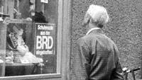 Ein Passant betrachtet die Auslagen im Schaufenster eines Schuhgeschäfts, im Fenster ein Schild: "Schuhe aus der BRD eingetroffen!"